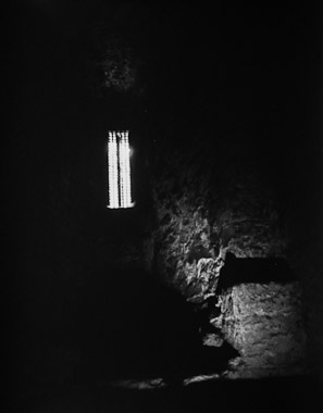 A black and white picture of a dark interior cell at the Tower of London with some illumination coming through a small window.